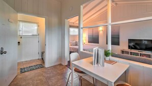 Una habitación compacta y bien iluminada, con cama, una mesita pequeña con una silla, televisor y ventana con persianas.