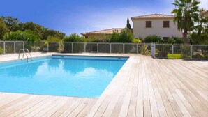 A rectangular swimming pool with a wooden deck surrounding it, a metal fence, and a building with a tiled roof in the background. - Appartement Lena. 6 pers,  piscine, 500 m plage (SAINTE LUCIE DE PORTO VECCHIO)
