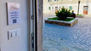 Eine Tür mit einem Schild, das Wohnungsnamen und einen QR-Code auflistet, ein gepflasterter Innenhof mit einem kleinen Busch und ein Gebäude mit Fenstern und Fensterläden im Hintergrund.