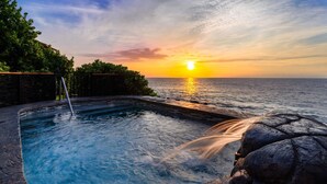 Ein Pool mit Wasserfall, umgeben von Grünflächen und mit Blick auf den Ozean bei Sonnenuntergang.