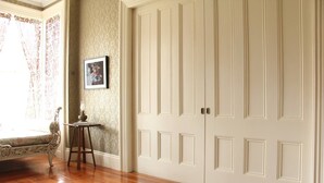 A room with a wooden floor, a white door with paneling, a framed picture on the wall, and a small table with a lamp. - The Wedding Cake House (Providence)