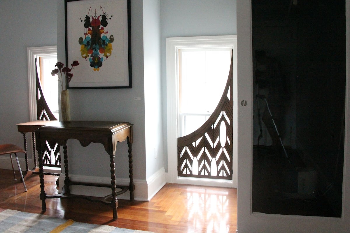 A room with a wooden floor, a small table, a framed abstract painting, and a window with a decorative screen.