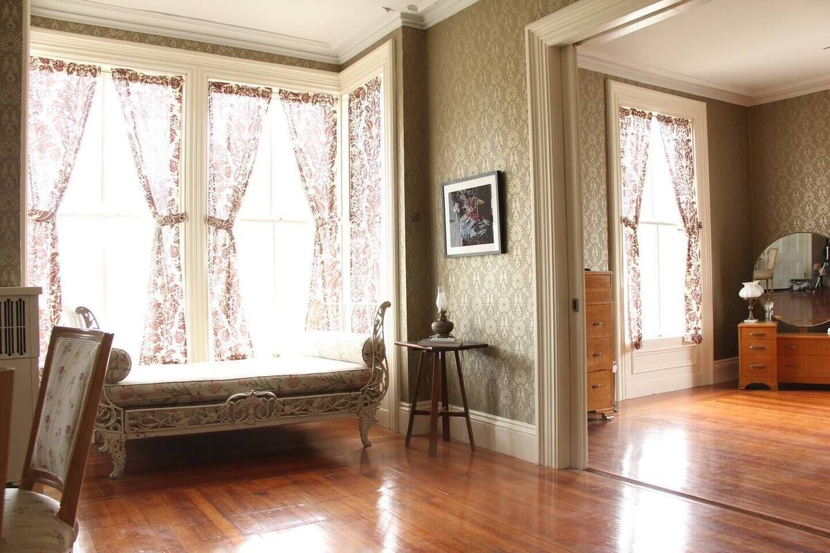 A room with a bay window, a wooden floor, a vintage bench, a small table with a lamp, and a dresser.