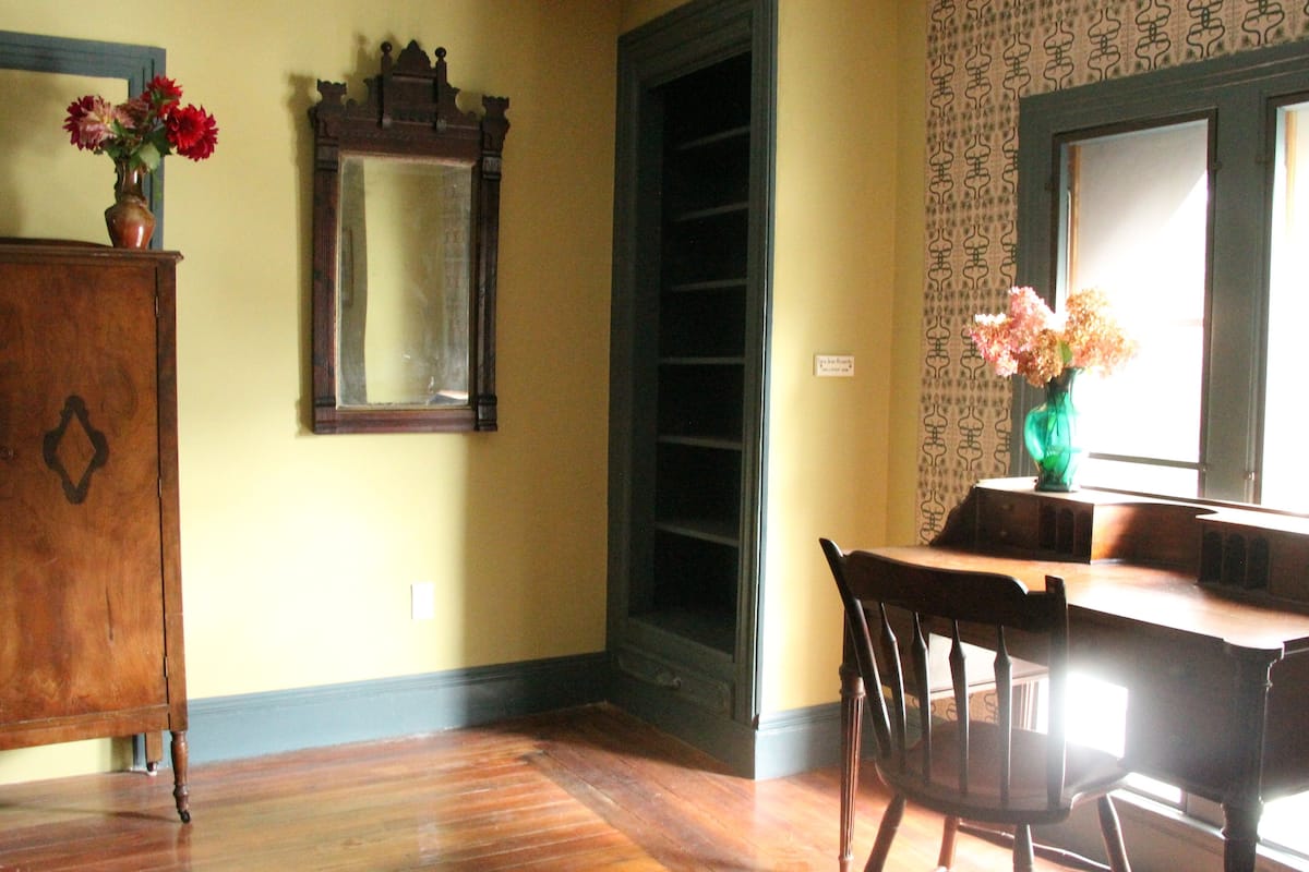 A room with wooden flooring, a wooden chair, a table, a mirror, and a window with patterned curtains.