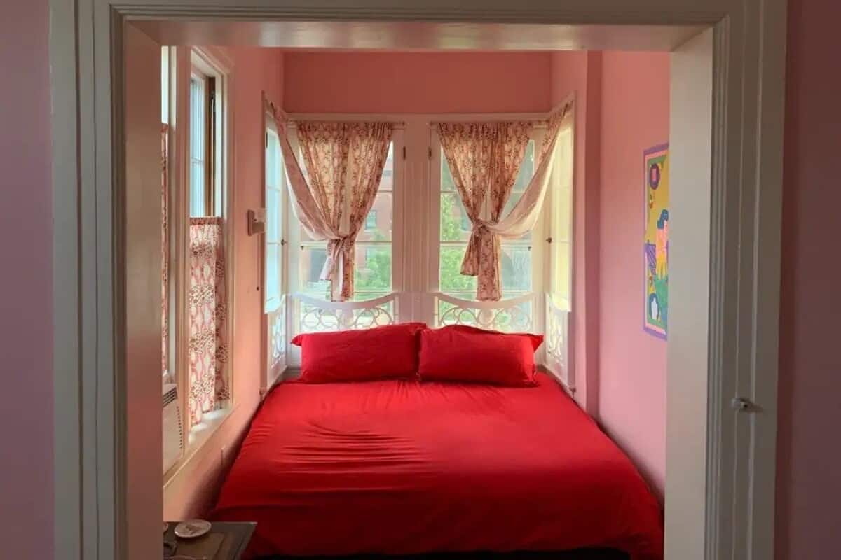 A bedroom with a red bedspread, two windows with curtains, and a door leading to another room.