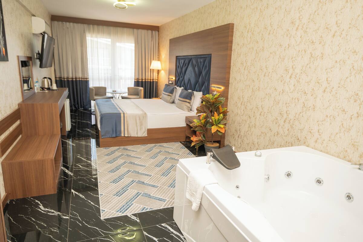 A hotel room with a bed, a sitting area with a chair, a desk, a TV, a bathroom with a bathtub, and a plant.