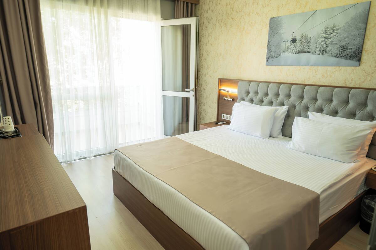 A hotel room with a large bed, a wooden desk, a nightstand with a lamp, and a painting on the wall.