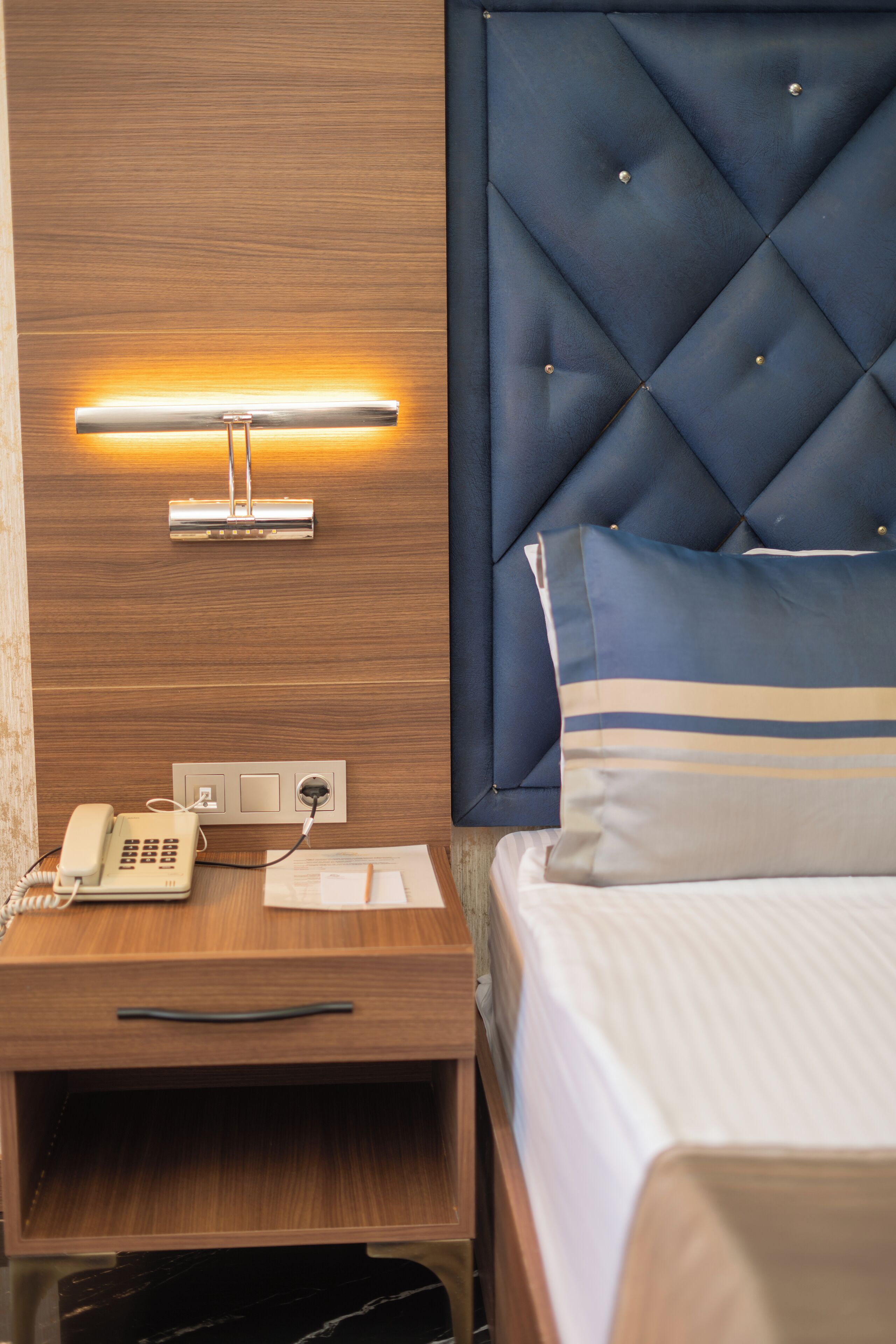 Una habitación de hotel con una mesita de noche de madera, un teléfono, una lámpara de pared, una cama con cabecera acolchada y una almohada a rayas.
