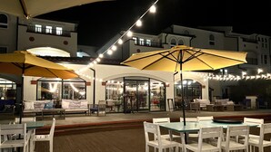 An outdoor patio area with white tables and chairs, yellow umbrellas, and string lights. - Beachfront Capriani Resort, Private Balcony, Ocean Pool Views, Linens Towels (North Topsail Beach)