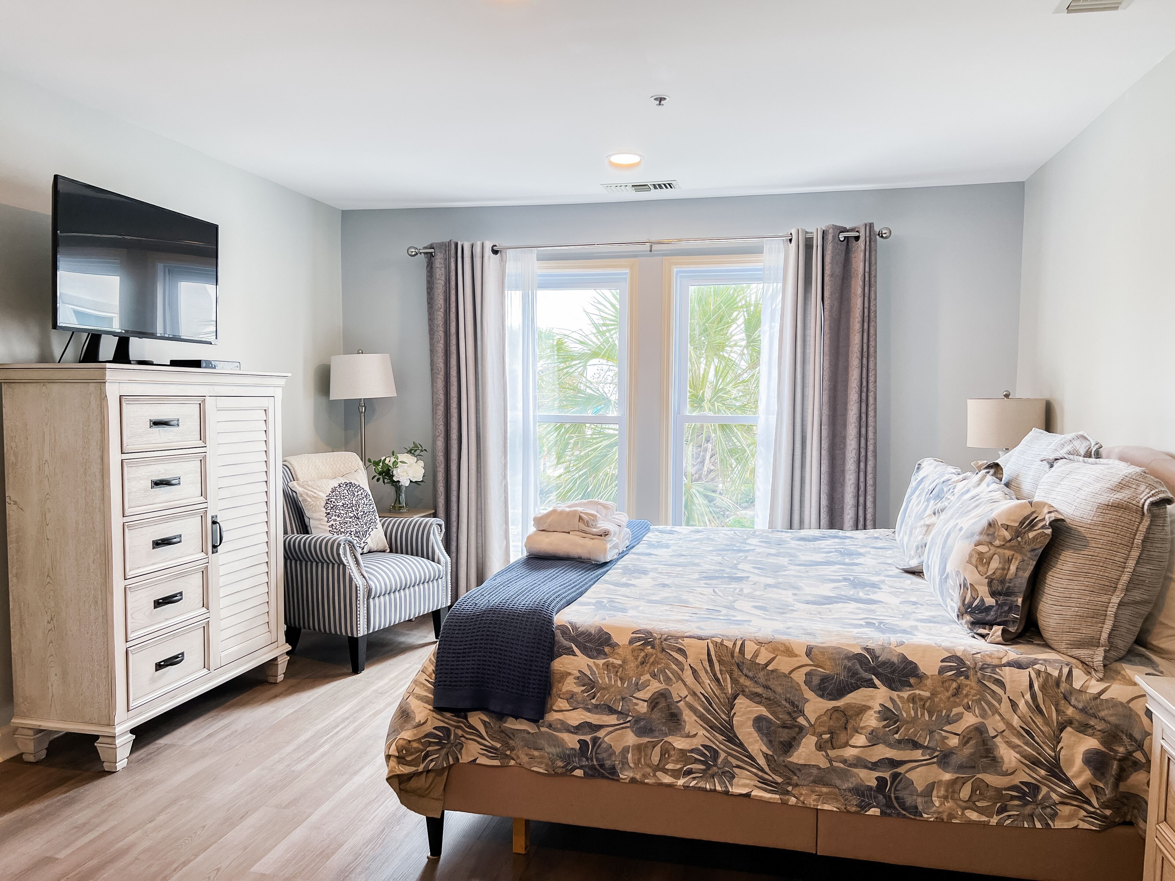 Een slaapkamer met een bed, een dressoir, een stoel, een televisie en een raam met gordijnen.