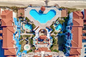 An aerial view of a resort with multiple swimming pools, a central courtyard, and surrounding buildings with red-tiled roofs. - Beachfront Capriani Resort, Private Balcony, Ocean Pool Views, Linens Towels (North Topsail Beach)