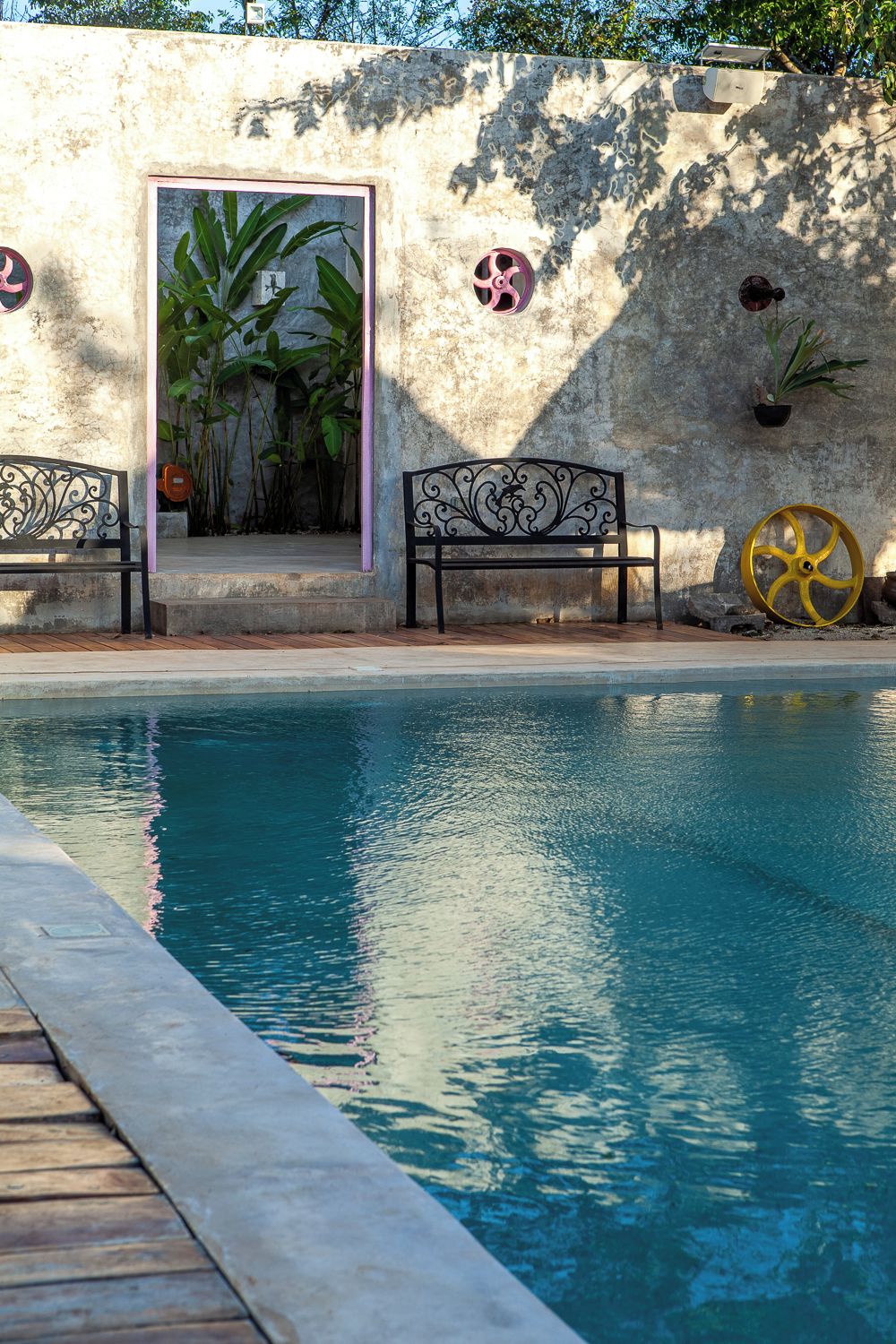 A swimming pool with a wooden deck, two ornate black benches, and a wall with a door and potted plants.