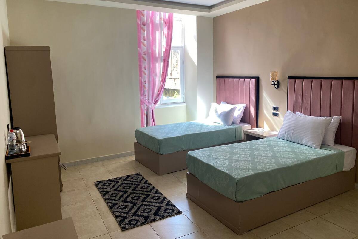 A hotel room with two beds, a small desk, a nightstand, and a window with pink curtains.