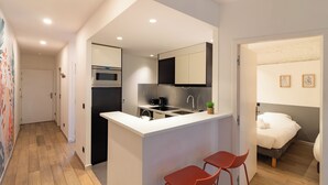 Una cocina compacta con una isla central, microondas y un lavabo. En la habitación contigua hay dos sillas rojas y una cama con ropa de cama blanca.