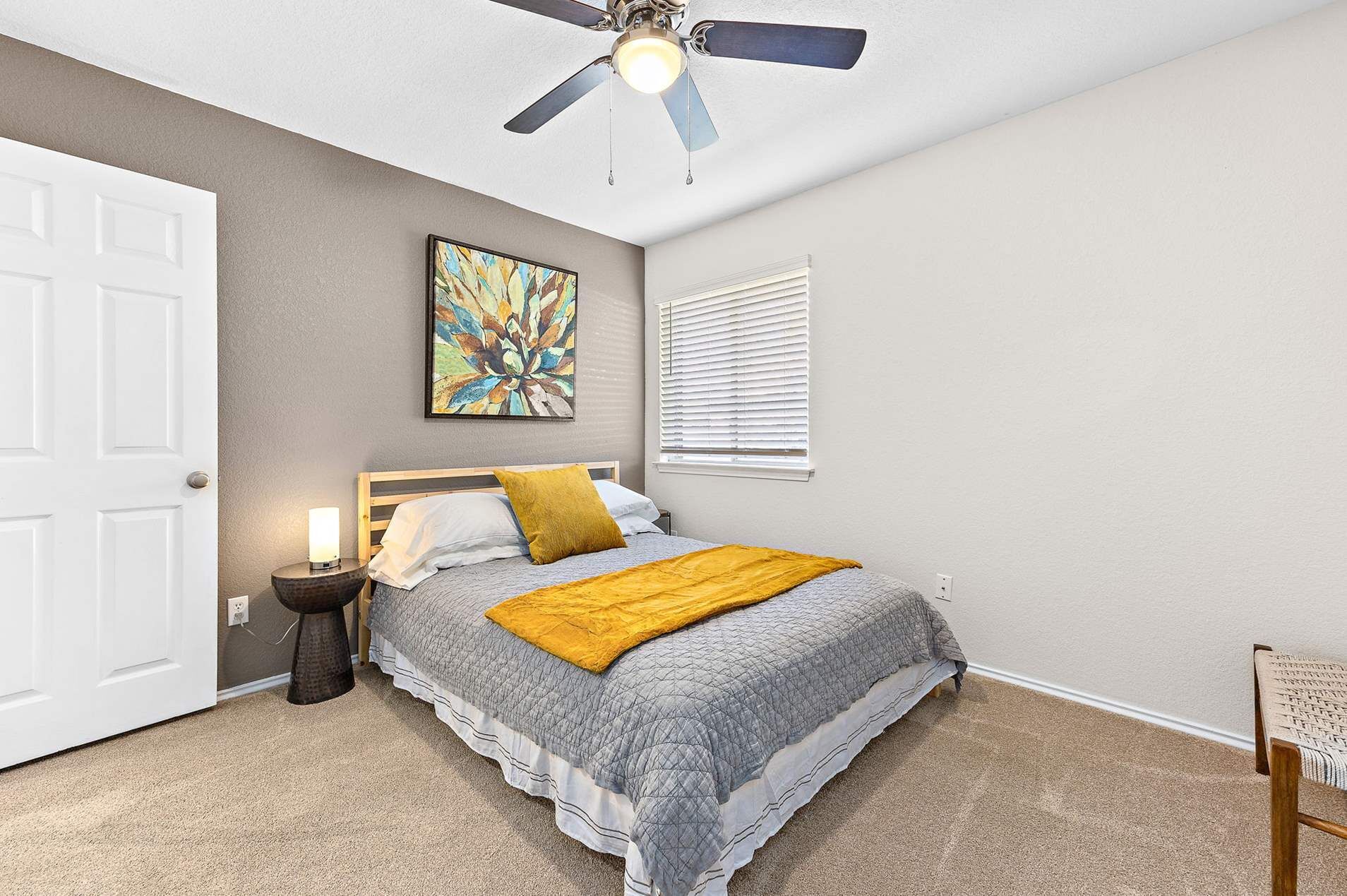 A bedroom with a bed, a ceiling fan, a window with blinds, a nightstand with a lamp, and a painting on the wall.