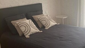 Une chambre à coucher avec une tête de lit sombre, deux oreillers à motifs circulaires, une petite table ronde et un interrupteur sur le mur.