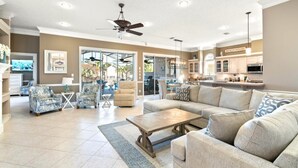 A spacious living room with a large sectional sofa, a coffee table, and a dining area with a table and chairs. There is a kitchen area with cabinets and a countertop visible.