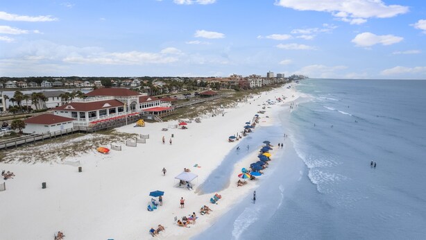 A beach with a long stretch of sand, umbrellas, and people. There are buildings and a pier in the background.