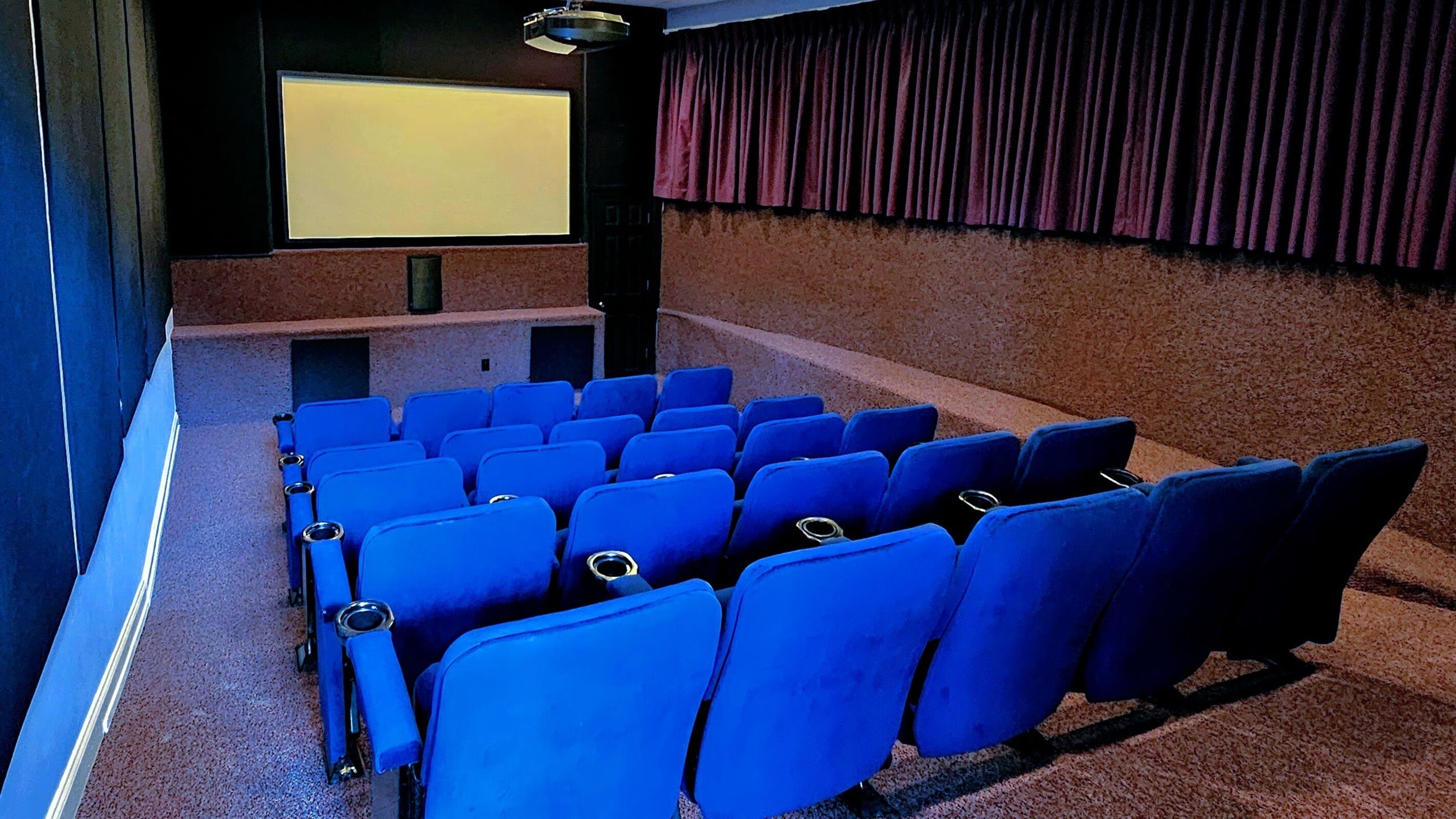 Ein Kino mit Reihen blauer Sitze, einer großen Leinwand und einem roten Vorhang im Hintergrund.