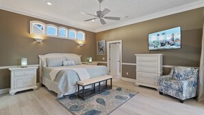 A bedroom with a bed, a bench, a dresser, a chair, a ceiling fan, and a wall-mounted TV.