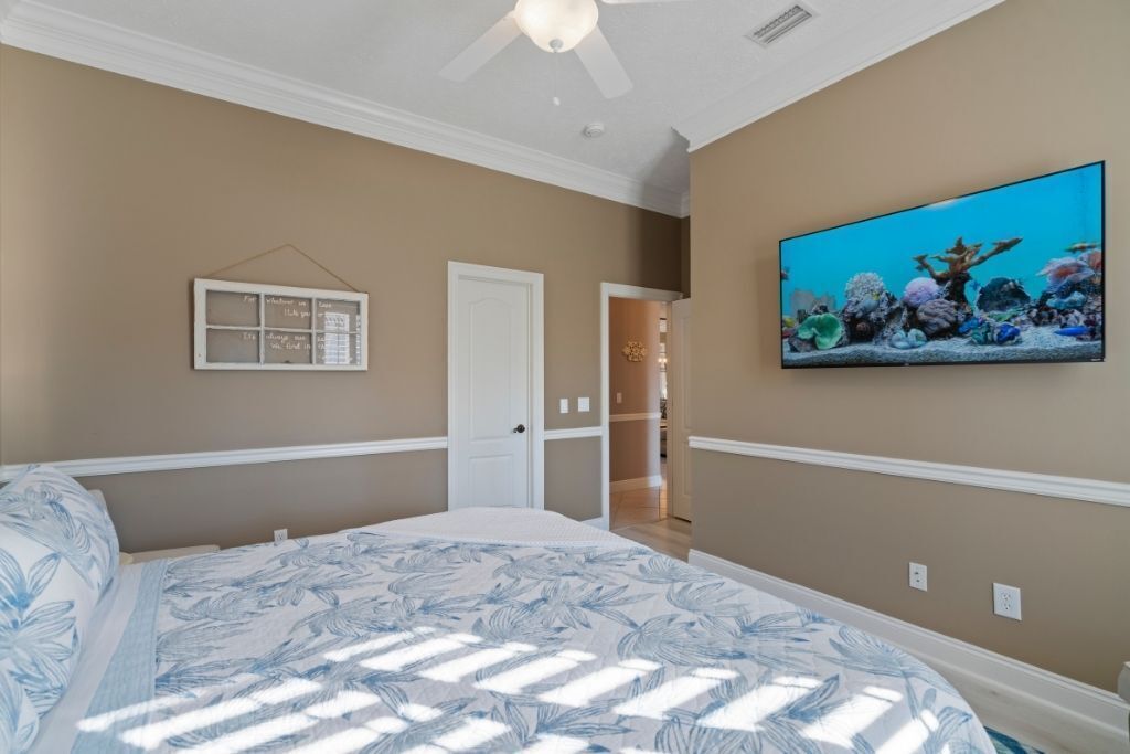 Un dormitorio con cama, ventilador de techo, televisor montado en la pared y una puerta que da a otra habitación.