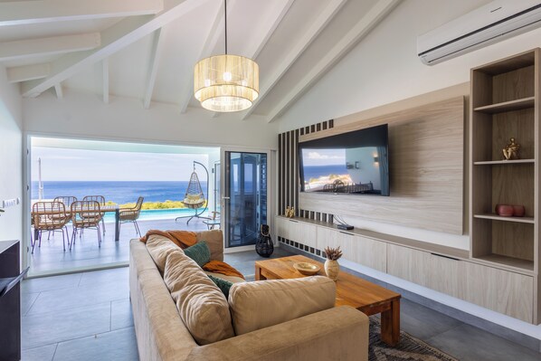 A modern living room with a large TV, a sofa, a dining area with chairs, and a view of the ocean. - Coral Estate Holidays (St. Marie)