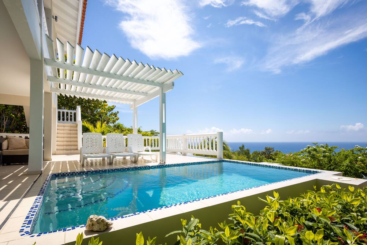 A pool with a tiled edge, surrounded by white lounge chairs and a pergola.