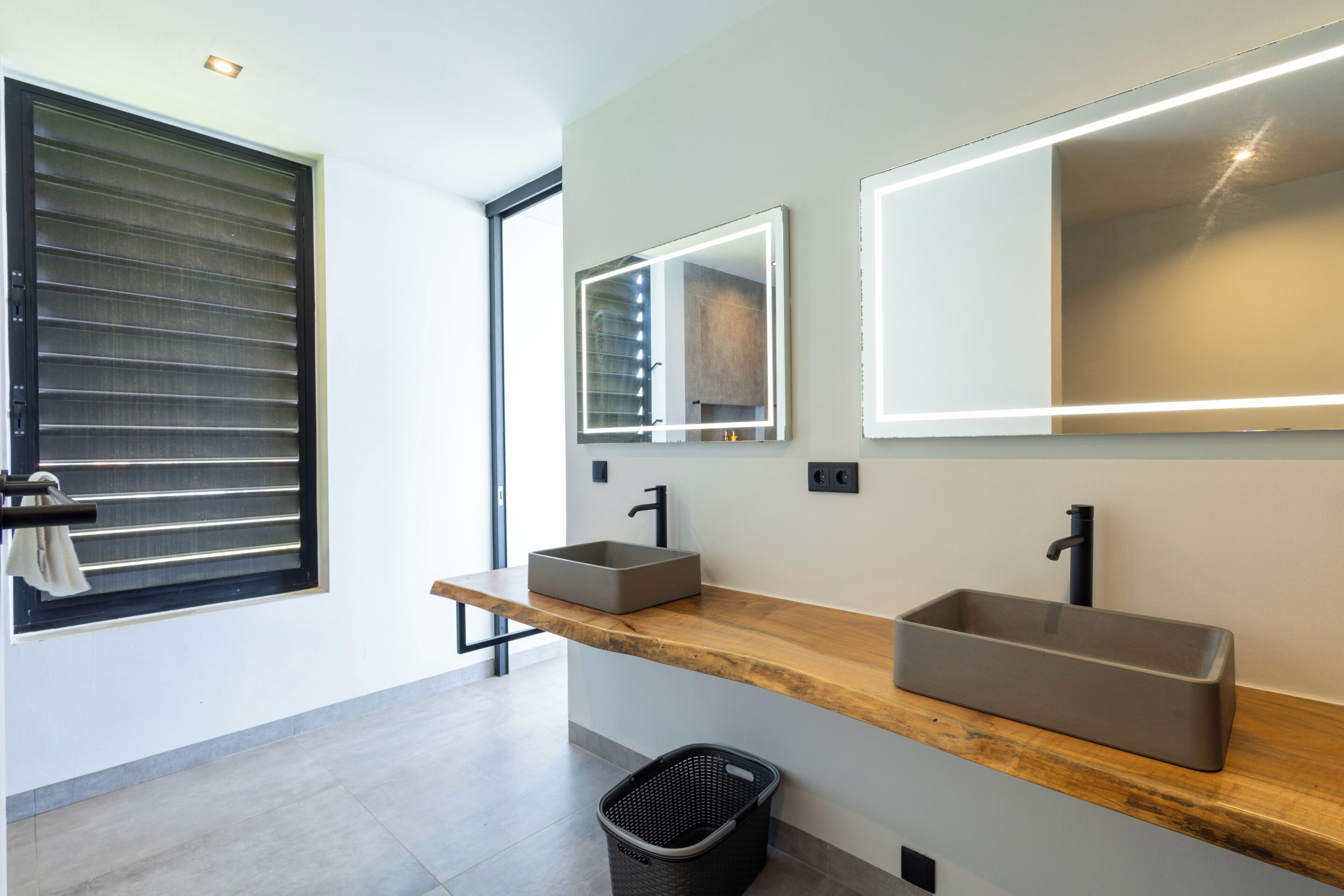 A modern bathroom with two sinks, a large mirror, and a window with blinds.