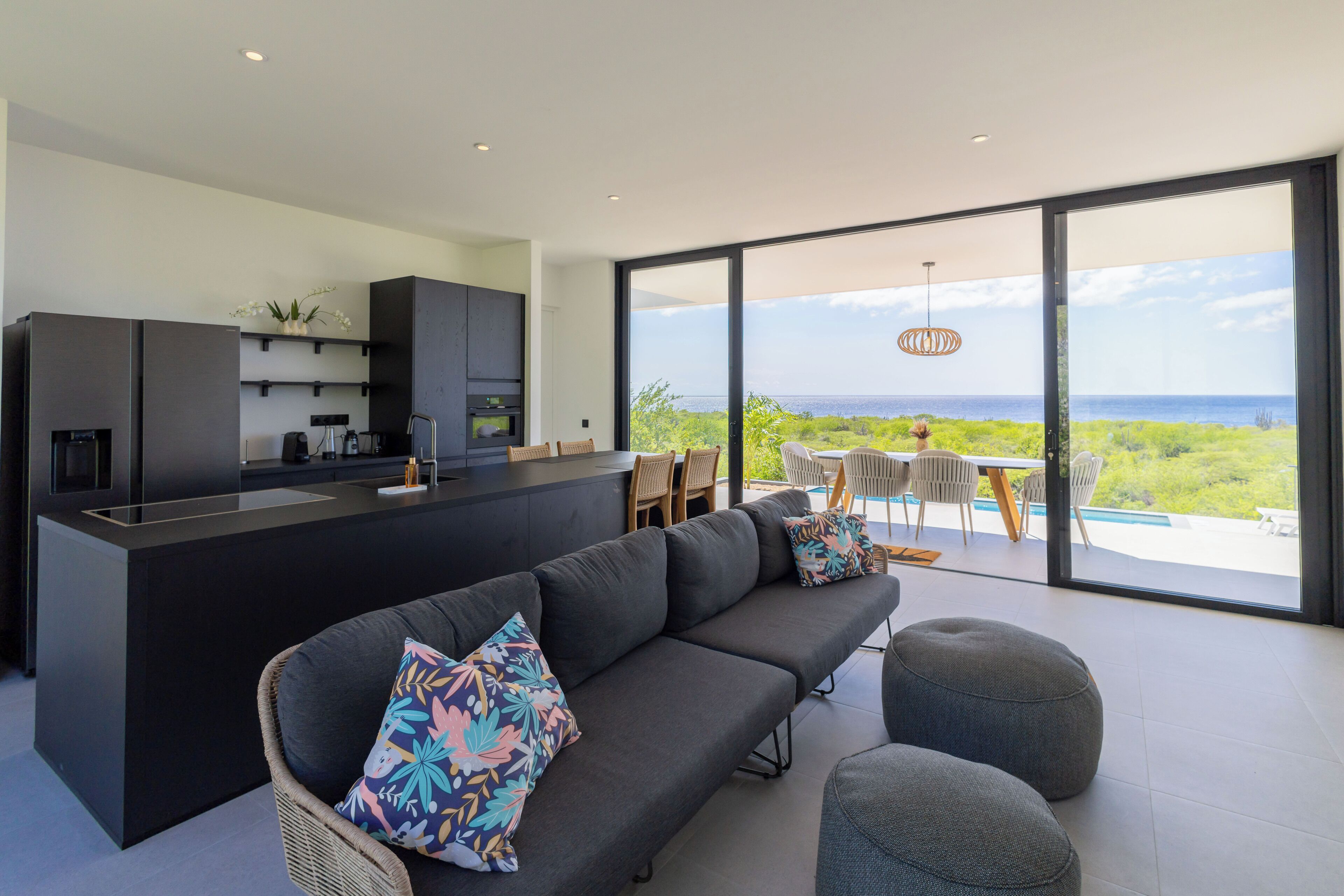 A modern kitchen with a central island, a dark sofa with colorful cushions, and a dining area with a view of the ocean.