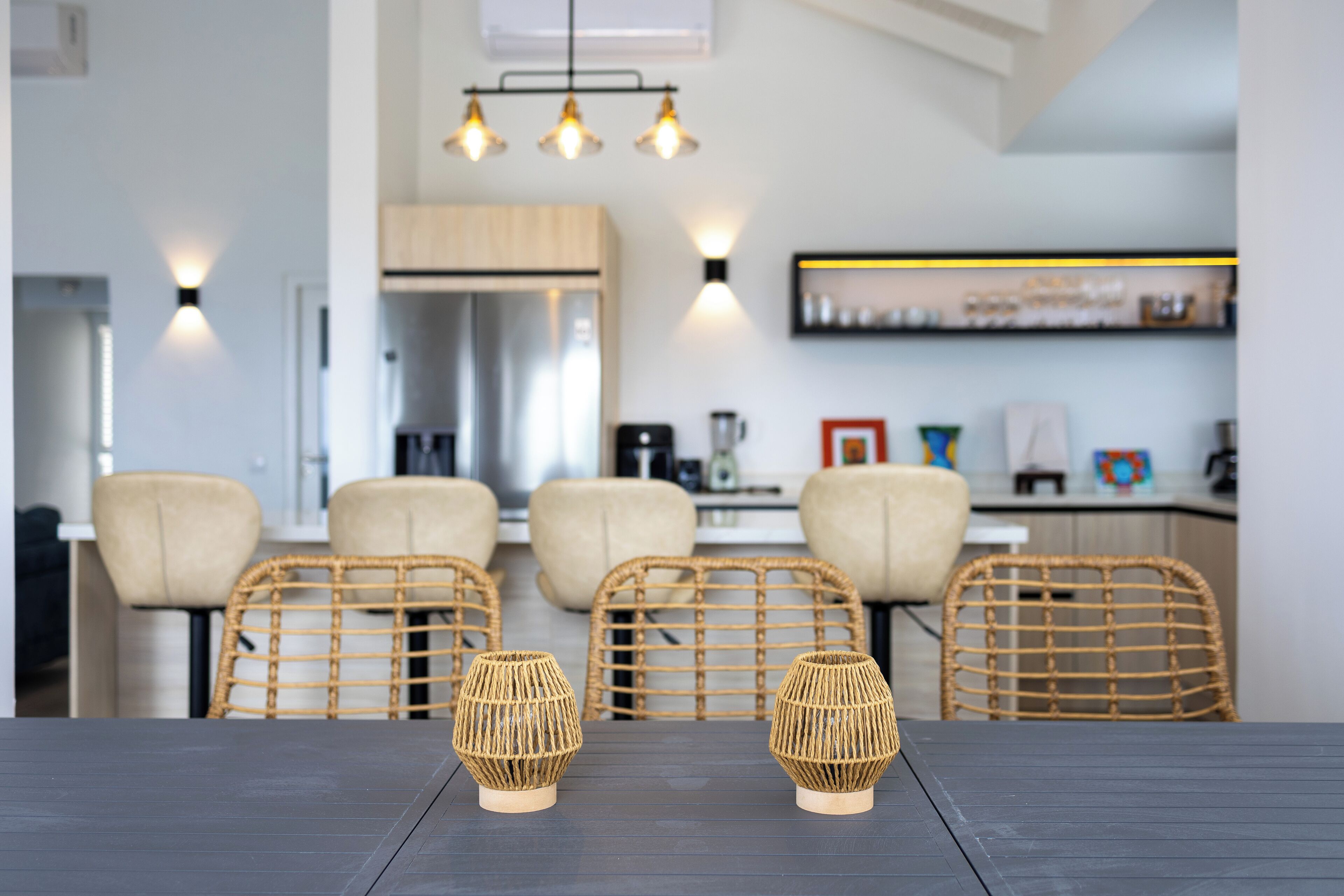 Une cuisine moderne avec un coin repas comprenant une table en bois foncé, des chaises en osier et deux photophores en fibres tressées.