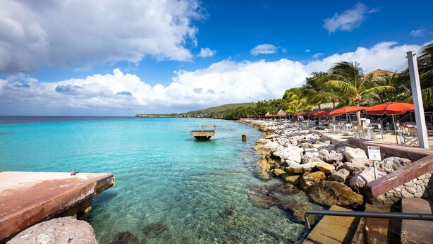A beach with clear turquoise water, a wooden boat, rocky shoreline, and red umbrellas. - Coral Estate Holidays (St. Marie)