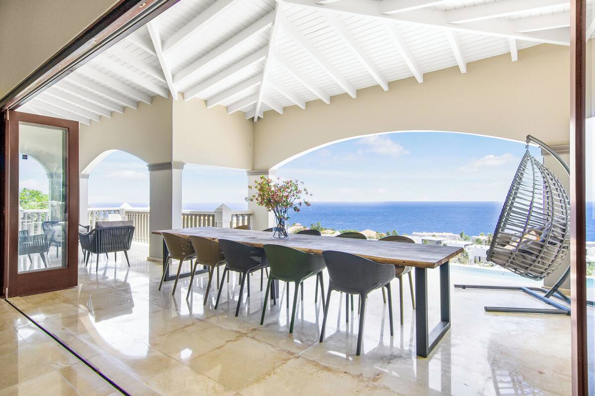 A covered outdoor dining area with a view of the ocean, a pool, and a hanging chair.