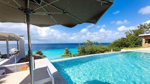 A clear blue pool with a view of the ocean, surrounded by greenery and a covered seating area. - Coral Estate Holidays (St. Marie)