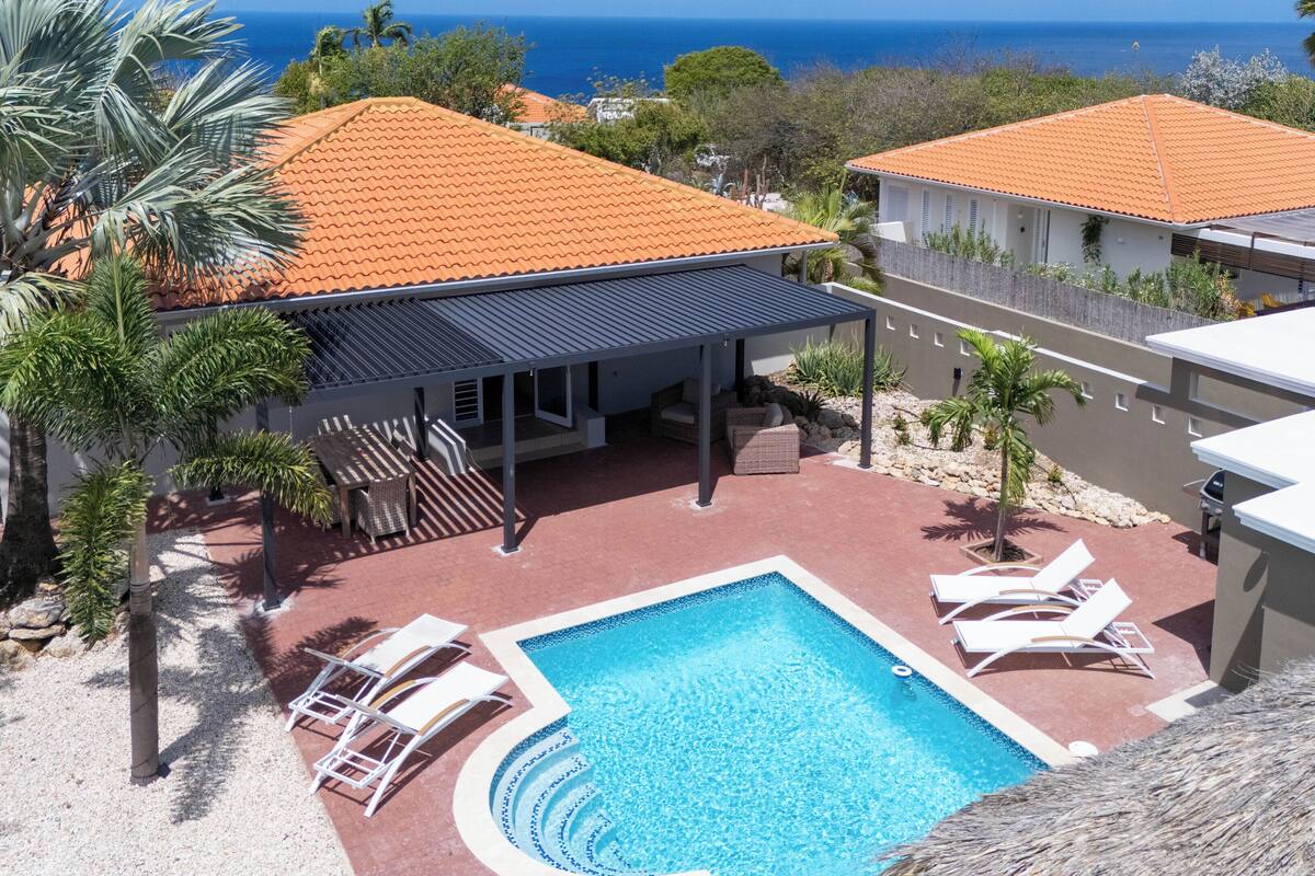 A pool area with a clear view of the ocean, a covered patio, and a building with a tiled roof.