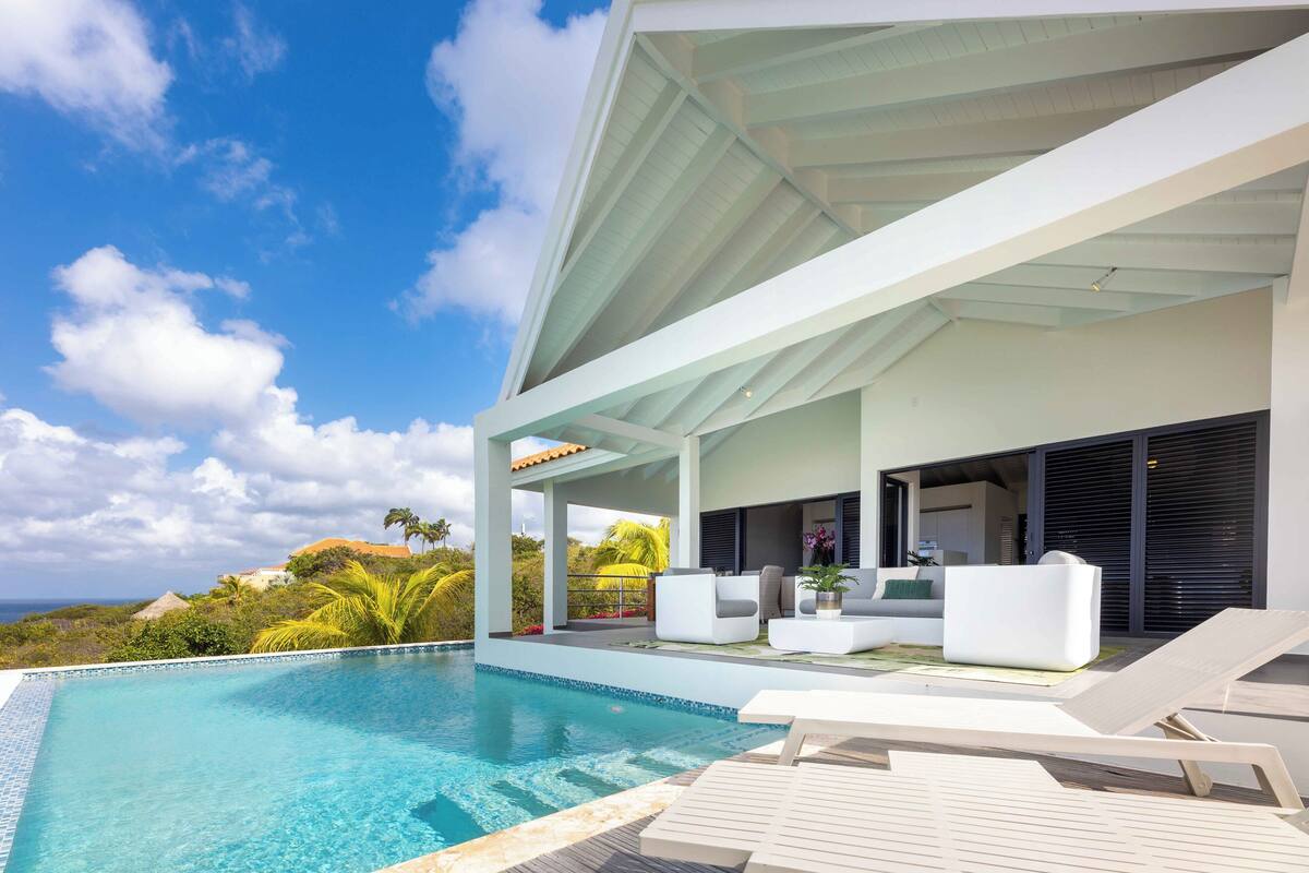 A modern outdoor pool area with lounge chairs, a covered seating area, and a view of the ocean.