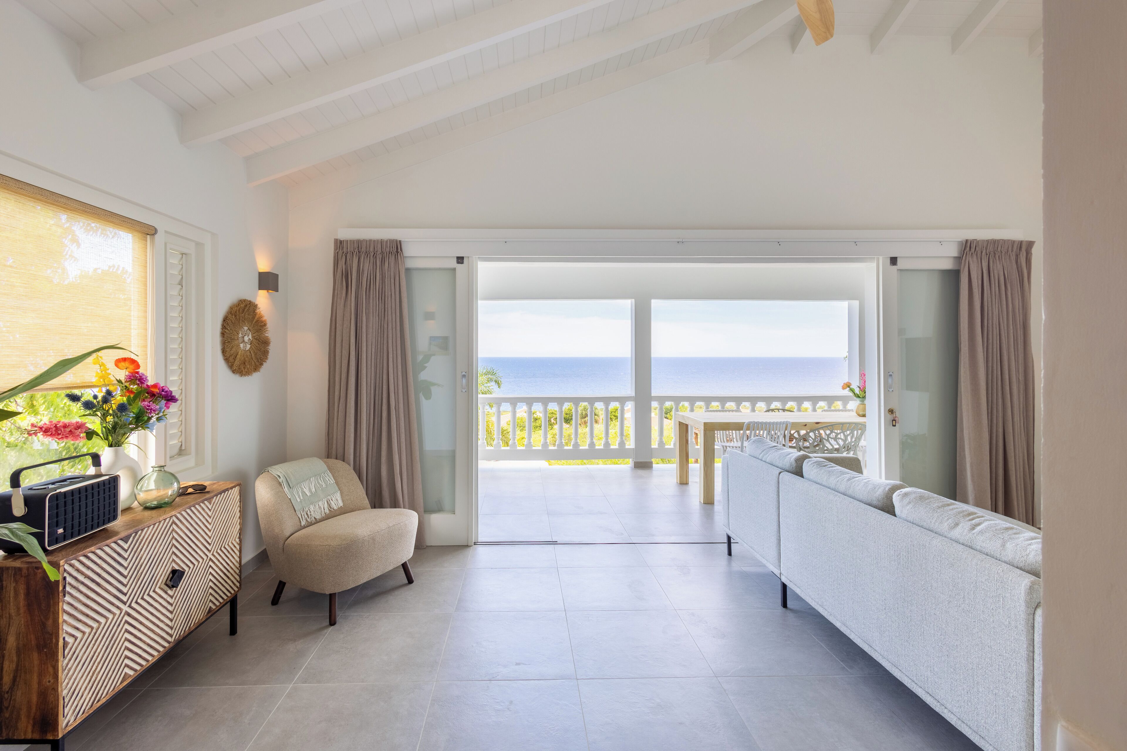 A spacious living room with a sofa, a chair, a wooden console table, and a view of the sea through large glass doors.