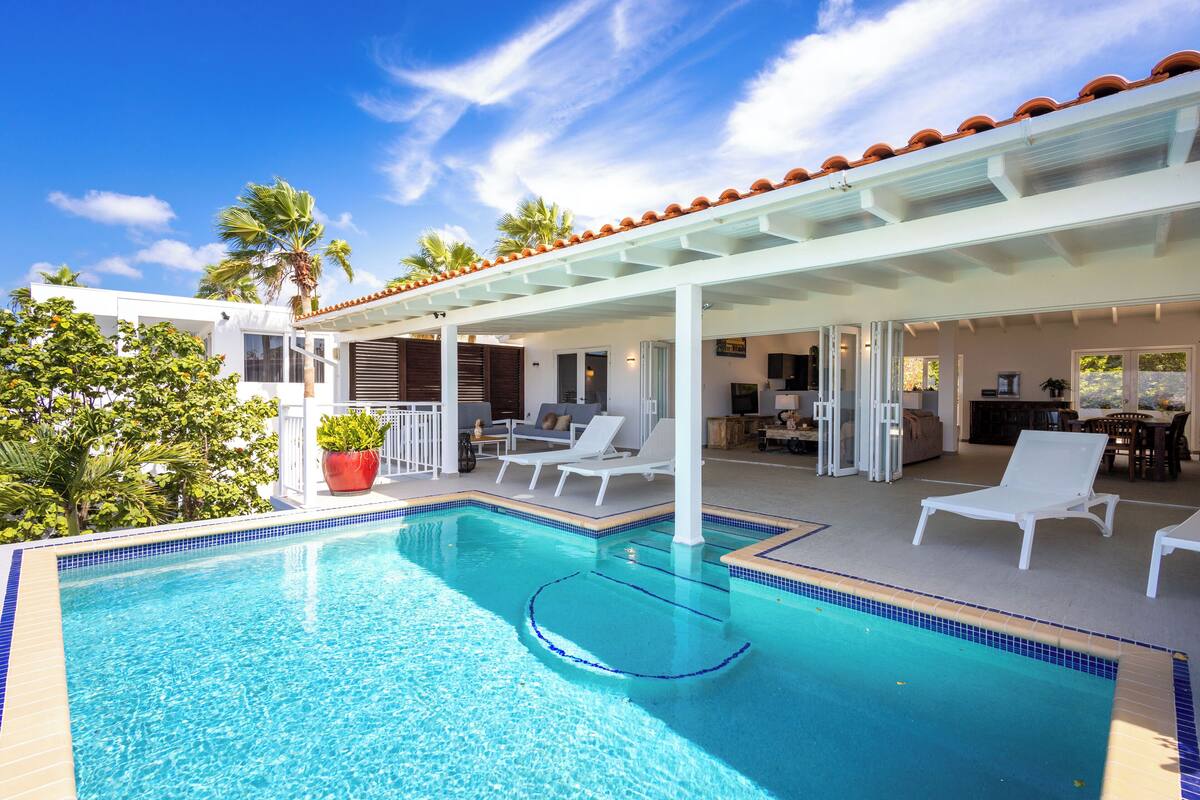 A swimming pool with lounge chairs, a covered patio, and a view of a house with a tiled roof.