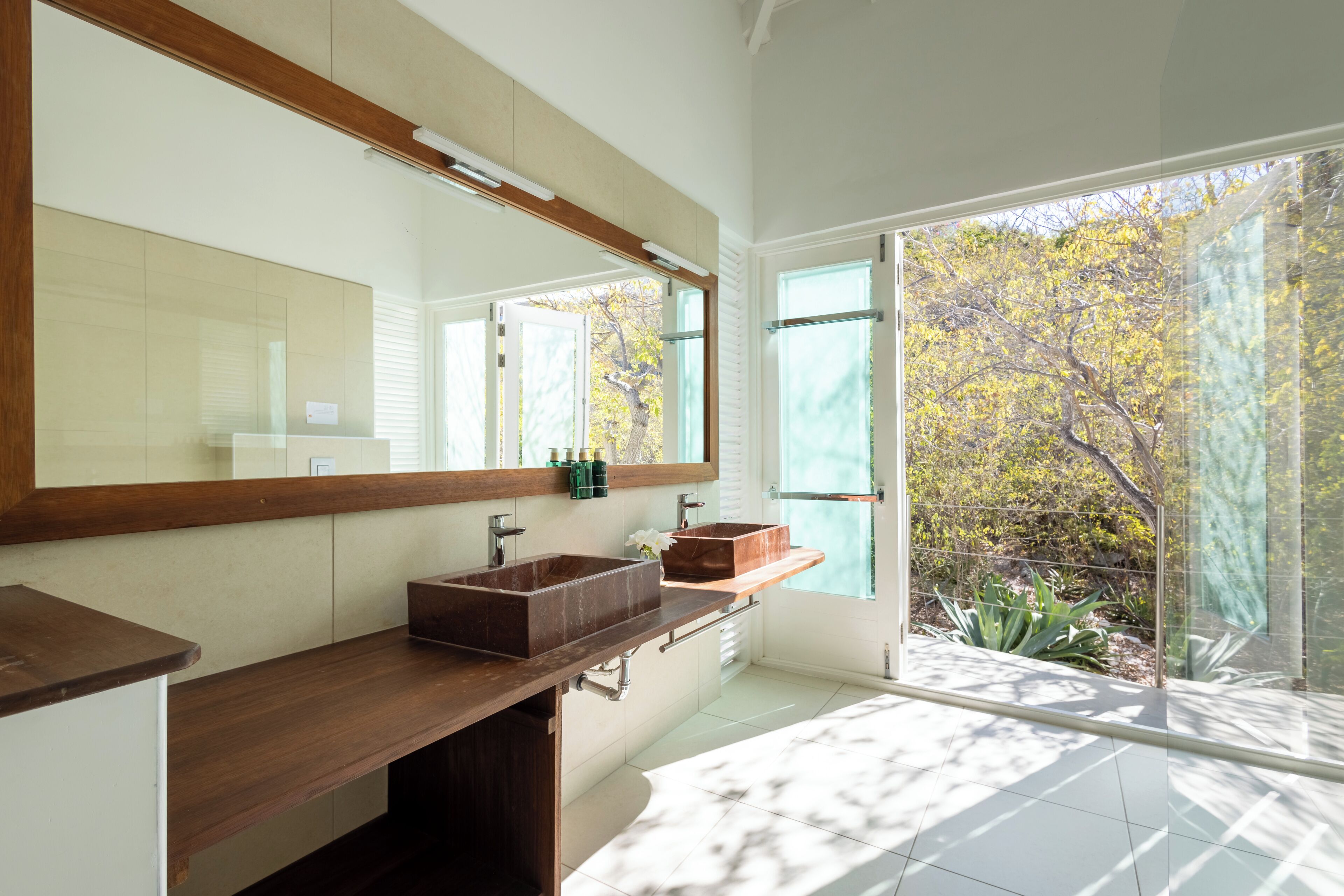 A modern bathroom with a large mirror, two sinks, and a view of trees through large windows.