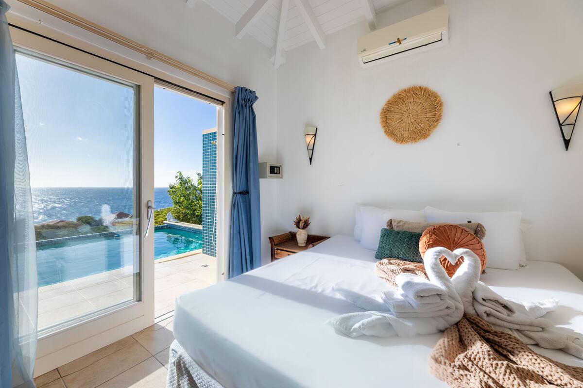 A hotel room with a bed, a view of the ocean, a pool, and a balcony.