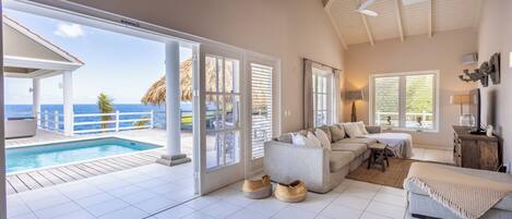 A spacious living area with a large window overlooking a pool and the ocean, a cozy seating area, and a ceiling fan.