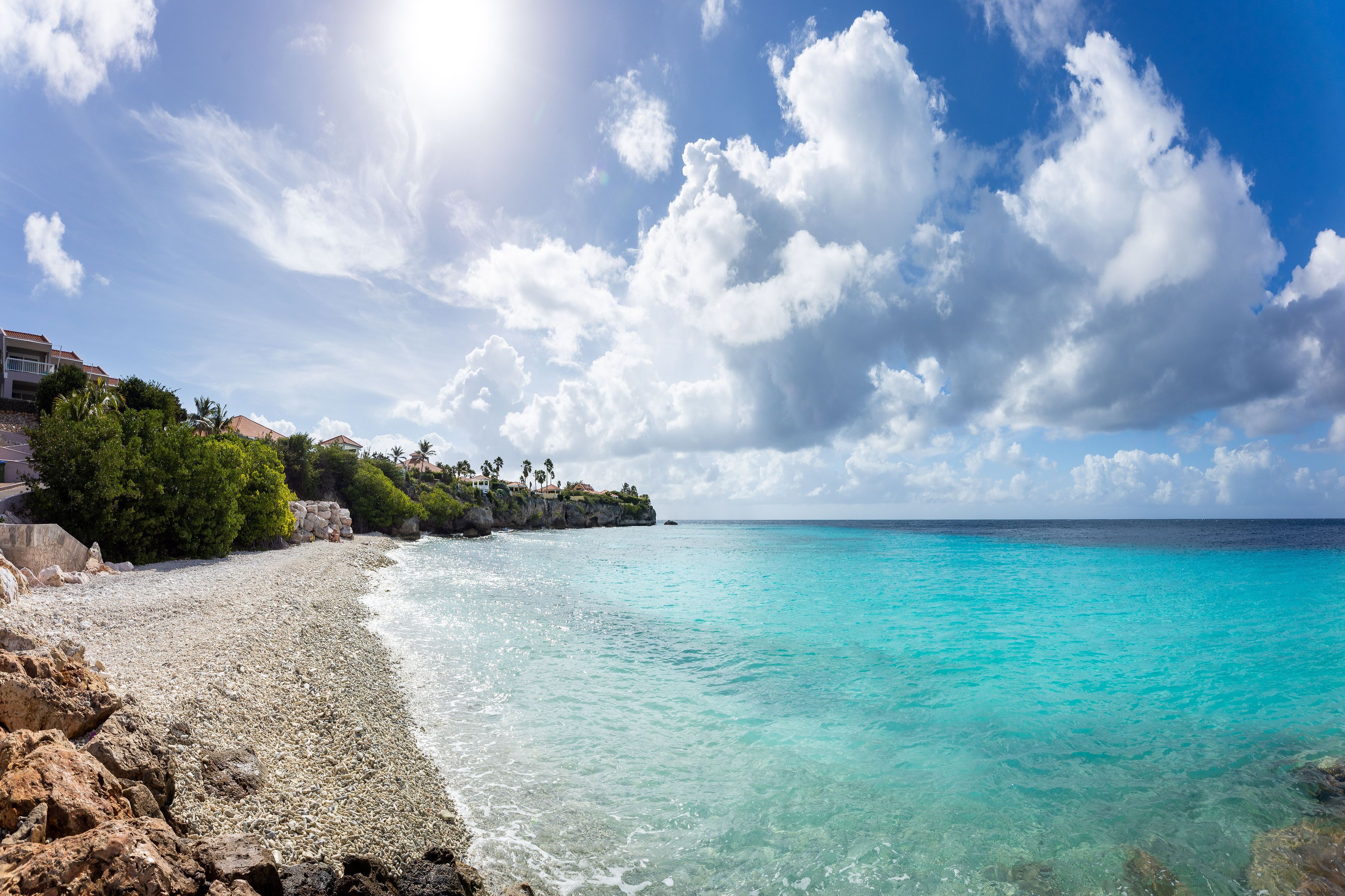 A beach with clear turquoise water, rocky shoreline, and a building with a balcony overlooking the sea.