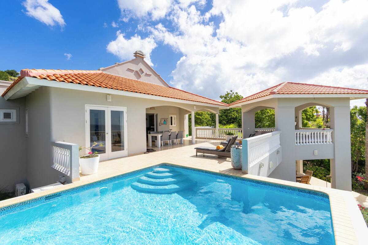 A swimming pool with a tiled deck, a covered patio area, and a white exterior wall.