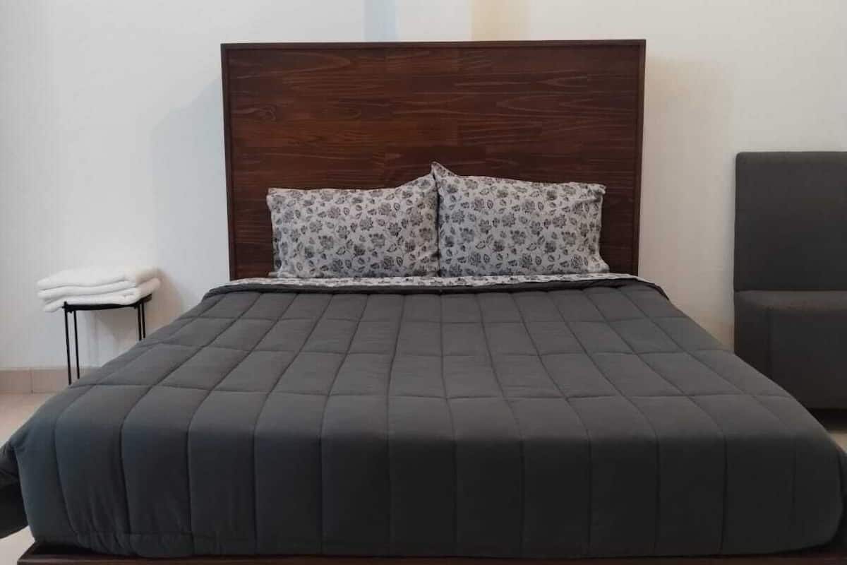 A bed with a dark headboard and two patterned pillows, a small bedside table with a folded white towel, and a chair to the right.
