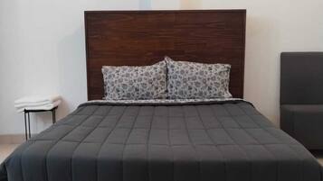 Un lit avec une tête de lit sombre et deux coussins à motifs, une petite table de chevet sur laquelle est pliée une serviette blanche, et une chaise sur la droite.