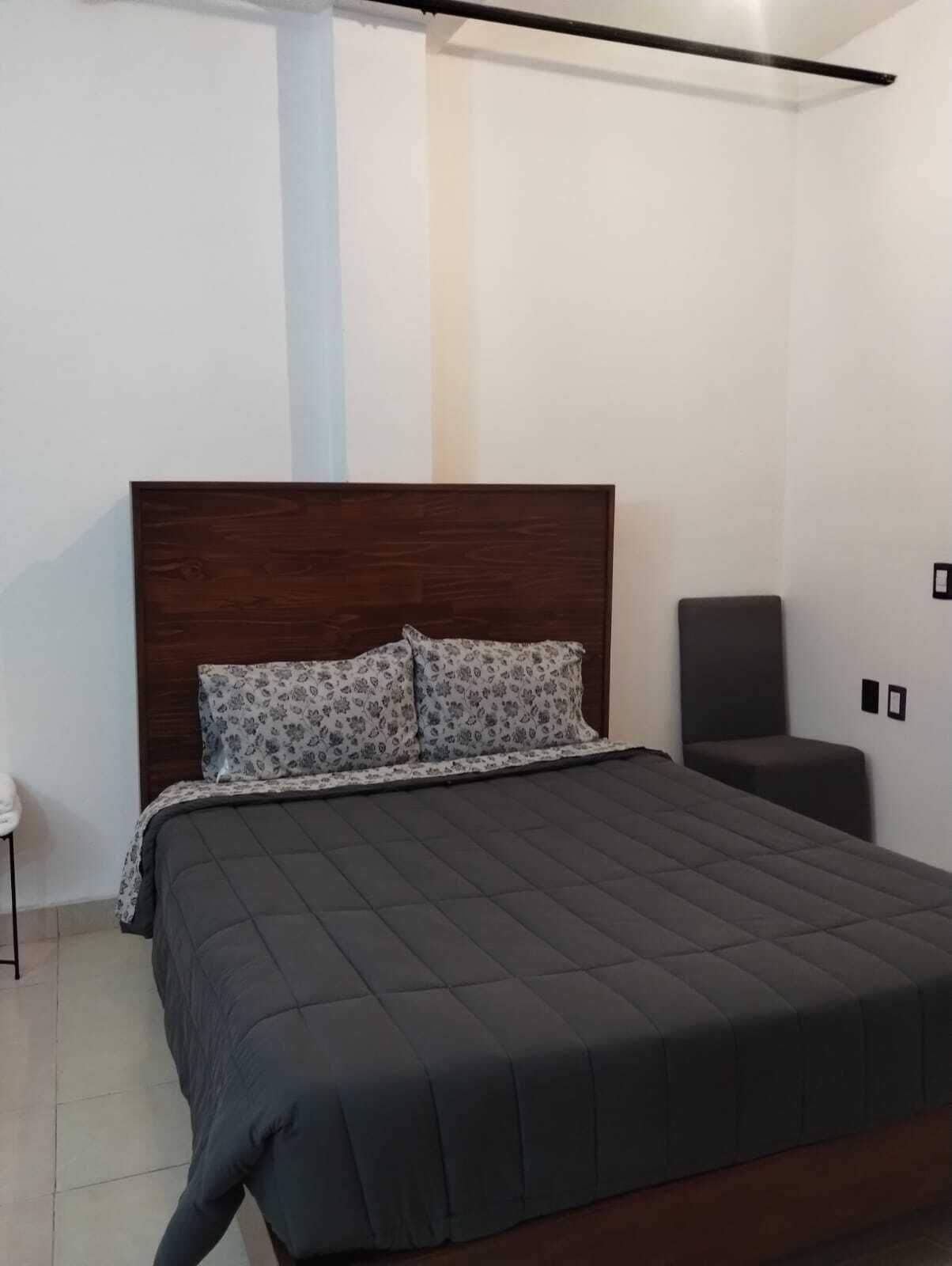 A bedroom with a dark wooden headboard, a bed with a grey quilt, two patterned pillows, a bedside table, and a chair.