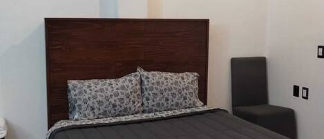 A bedroom with a dark wooden headboard, a bed with a grey quilt, two patterned pillows, a bedside table, and a chair.