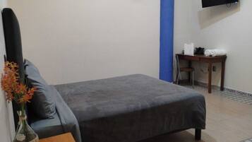 Habitación de hotel con cama, escritorio, silla, televisor montado en la pared y un florero con flores sobre la mesita de noche.