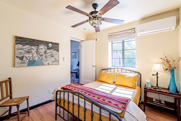 A bedroom with a bed, a ceiling fan, a window, a nightstand, a lamp, and a painting on the wall.