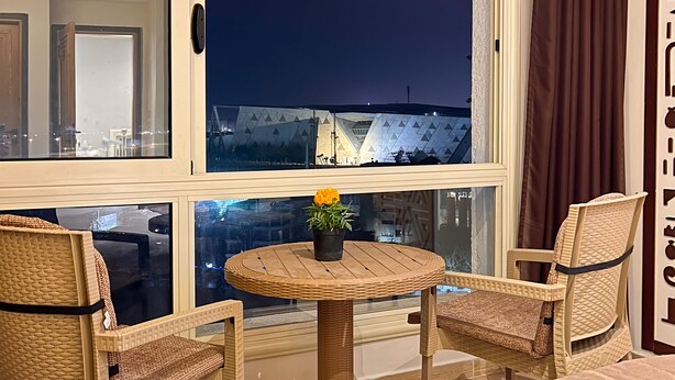 A hotel room with a large window, wicker chairs, a small round table with a flower, and a bed. - Grand Museum Guest House pyramids view (Giza)