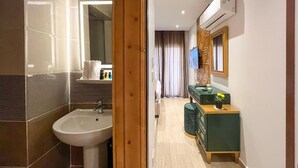 A modern bathroom with a sink and mirror, a wooden door, and a hallway leading to a room with a green dresser and a TV. - Grand Museum Guest House pyramids view (Giza)
